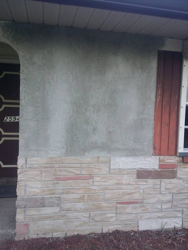 Blotchy weathered stucco above a stone veneer band, beside red window shutters